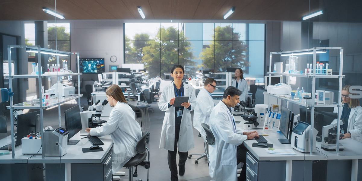 Modernes Forschungslabor mit mehreren Wissenschaftlern in weißen Laborkitteln, die an Mikroskopen und Computern arbeiten, während eine Forscherin mit Tablet im Vordergrund steht.